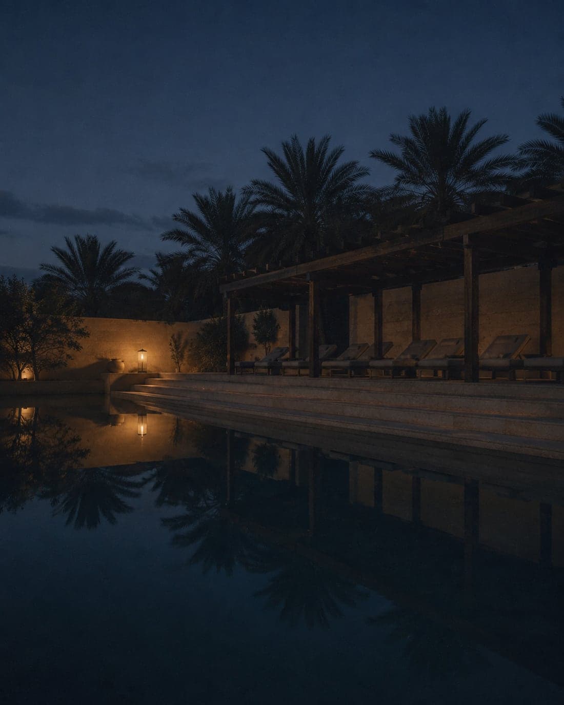 A still pool at night beside a timber pergola of empty loungers, palms and one lantern reflected in the water