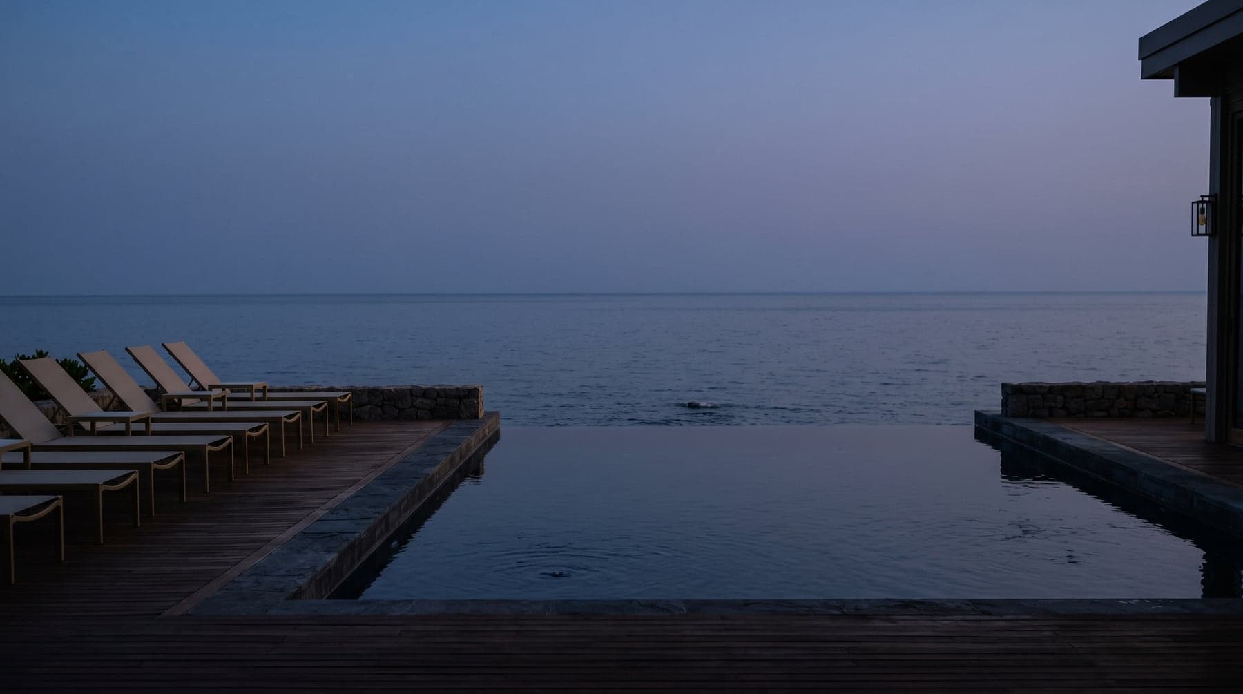 An infinity pool meeting a calm sea at blue hour, empty loungers along the timber deck