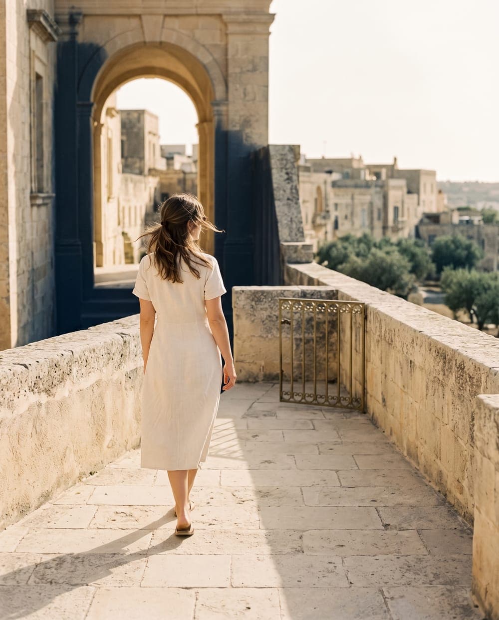 A woman in a cream linen dress walking alone along a sunlit limestone parapet, an old hill town beyond